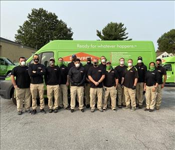 Group of men & women standing in uniforms and masks in front of SERVPRO trucks