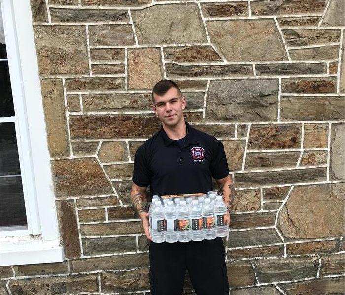 male firefighter standing against fire house with a case of SERVPRO of Newark water 